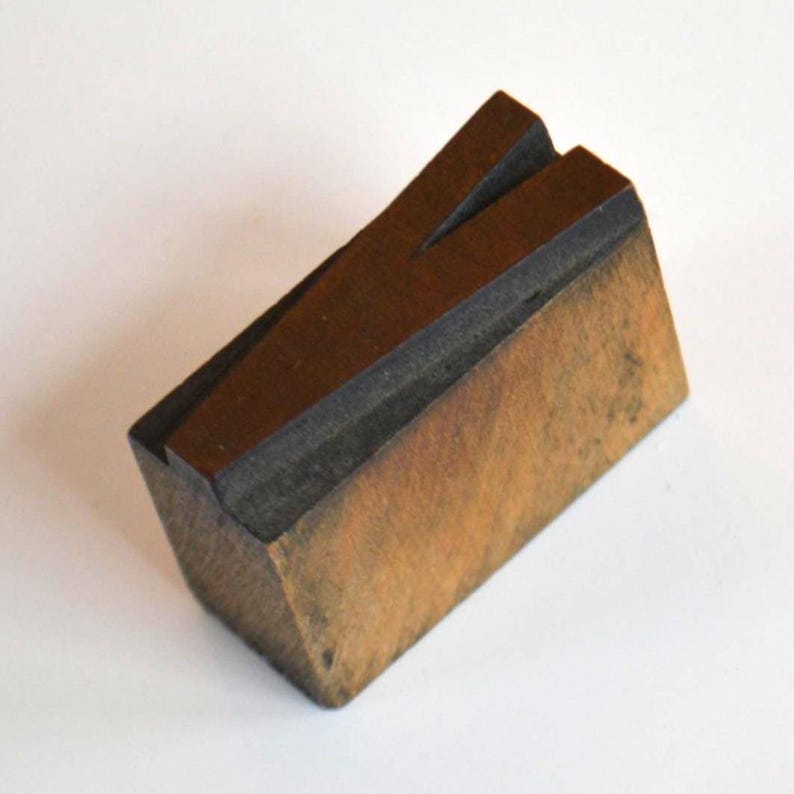 May include: A wooden letterpress printing block in the shape of the letter "V". The block is made of wood and has a dark brown printing surface. The block is angled and sits on a white surface.