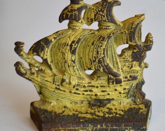 May include: A cast iron doorstop shaped like a sailing ship with multiple masts and sails. The object features a distressed yellow paint finish over a dark brown metal base, showing visible wear and texture across the hull and sails.