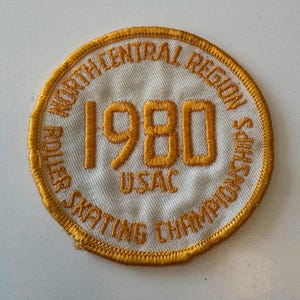 May include: A white, round patch with gold embroidered text. The text reads "North Central Region Roller Skating Championships 1980 USAC".