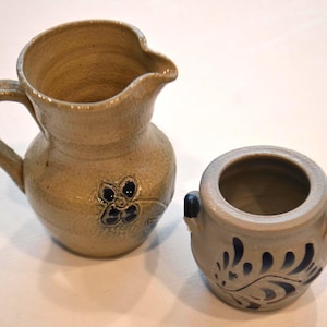 May include: Two ceramic pottery pieces, a pitcher and a small pot, both with blue and white floral designs. The pitcher has a handle and a wide mouth. The pot is round with a small opening.