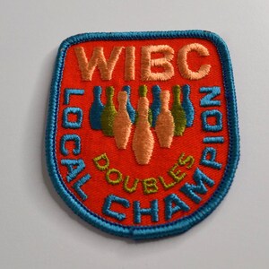 Könnte beinhalten: Ein schildförmiger, bestickter Aufnäher mit leuchtend orangefarbenem Hintergrund und blauem Rand. Er zeigt den Text WIBC LOCAL DOUBLES CHAMPION in Blau und Gelb, der eine zentrale Grafik von fünf Bowling-Pins in Pfirsich- und Grüntönen umgibt.