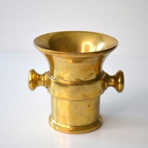 May include: A vintage brass mortar with a flared rim and two side handles. The mortar has a cylindrical base and a polished, golden finish. This apothecary tool is ideal for grinding herbs and spices.