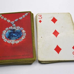 May include: Playing cards. One deck has a red back with a diamond pendant design. The other shows the three of diamonds. The cards are slightly worn.