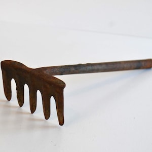 May include: A small, rusty metal garden rake with five tines and a long handle. The tool is made of weathered metal, showing signs of age and use. The rake is designed for gardening and landscaping tasks.