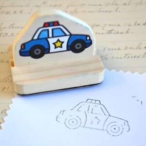 May include: A wooden stamp featuring a blue and white police car with a yellow star on the side and red and blue lights on top. The stamp has a matching outline imprint on a white paper.