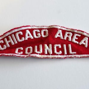 Puede incluir: Un parche rojo arqueado con texto bordado en blanco que dice "CHICAGO AREA COUNCIL". El parche tiene un borde blanco y probablemente sea un artículo vintage o de colección. Sobre un fondo blanco.