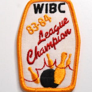 May include: A rectangular embroidered patch with rounded corners and an orange border. The white background features the black text WIBC, yellow text 83-84, and red cursive text League Champion above an illustration of a black bowling ball and pins.
