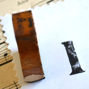 May include: A vintage wooden stamp with the letter "I" printed in black ink on white paper. The stamp has a rectangular shape with a brown, aged appearance. The background includes sheet music and a decorative paper edge.