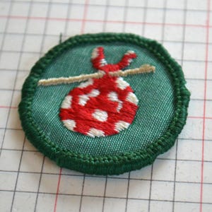 May include: A circular embroidered patch featuring a teal green background with a dark green stitched border. The center displays a red bundle with white polka dots, tied to a light tan stick, creating a classic hobo-style sack design.