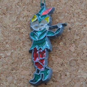 May include: A green and red glass ornament depicting a whimsical elf playing a flute. The elf has a green hat, green and red clothing, and yellow hair. The ornament is made of glass and has a silver finish.