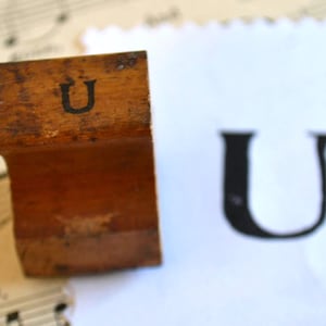 Könnte beinhalten: Ein Holzstempel mit dem Buchstaben "U" in schwarzer Farbe. Der Stempel ist braun und weist Gebrauchsspuren auf. Ein gedrucktes Bild des Buchstabens "U" befindet sich neben dem Stempel auf weißem Hintergrund. Der Hintergrund enthält Notenblätter.