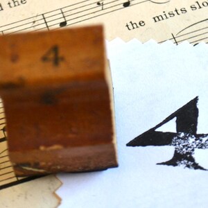 Könnte beinhalten: Ein Holzstempel mit der Zahl vier und einem schwarzen Tintenabdruck der Zahl vier auf weißem Papier. Der Stempel ist braun und trägt die Zahl vier. Der Hintergrund enthält Notenblätter.
