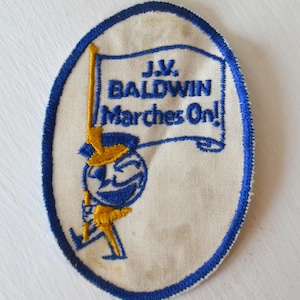 Può includere: Una toppa ovale bianca con cuciture blu intorno ai bordi. La toppa presenta un personaggio dei cartoni animati che tiene uno striscione con su scritto "J.V. Baldwin Marches On!" Il personaggio indossa un'uniforme blu e gialla e ha un viso sorridente.