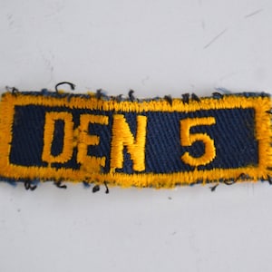 May include: A blue and yellow fabric patch with the text "DEN 5" embroidered in yellow.