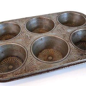 Puede incluir: Un molde para muffins vintage y usado con seis moldes circulares. La bandeja de metal tiene un aspecto envejecido y desgastado con patrones decorativos de estrellas alrededor de cada taza y en los bordes exteriores. Dos pequeños agujeros son visibles en el lateral.