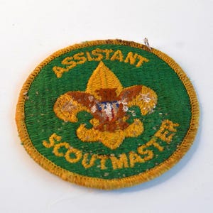 May include: A circular, green embroidered patch with the words "ASSISTANT SCOUTMASTER" in yellow. The patch features a gold fleur-de-lis emblem with brown and blue accents, and a gold border.