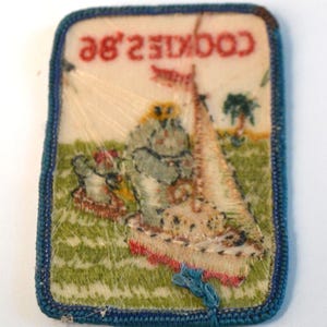 May include: A rectangular patch with a blue border and the text "COOKIE 2.8E" in red. The patch features a sailboat, a tree, and a figure on a green background. The patch is likely a vintage collectible.