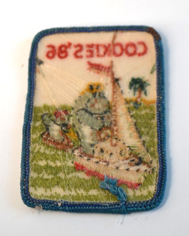May include: A rectangular patch with a blue border and the text "COOKIE 2.8E" in red. The patch features a sailboat, a tree, and a figure on a green background. The patch is likely a vintage collectible.