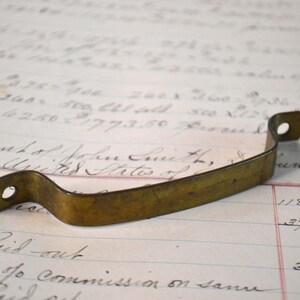 May include: A curved brass drawer pull with a weathered, aged patina. The handle features a flat, elongated center section that arches upward, ending in two flat, circular mounting tabs, each containing a single pre-drilled hole for attachment.