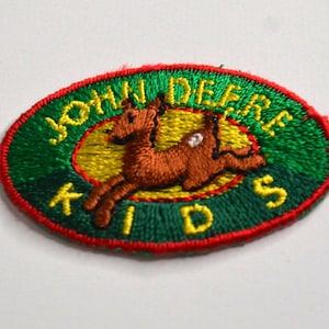 May include: An oval-shaped embroidered patch featuring a green background with a red border. The text JOHN DEERE KIDS is stitched in yellow thread. A brown deer is depicted in the center against a yellow oval background.