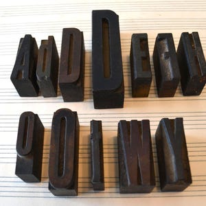 May include: A selection of antique wooden letterpress printing blocks. The dark brown blocks are arranged on a sheet of lined paper. The letters include A, D, E, M, O, I, N, and V. These blocks are used for printing.