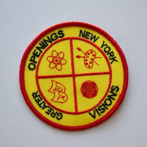 Peut inclure: Un patch circulaire jaune et rouge avec le texte "Greater New York Openings Visions" autour du bord. Le centre du patch est divisé en quatre quadrants avec différents symboles : un atome, un pinceau et une palette, un bâtiment et une pelote de laine.