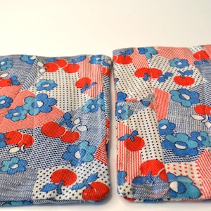 May include: Two square fabric items with a patchwork design. The fabric features a pattern of blue flowers, red cherries, and white polka dots on a blue and white background. The items are likely coasters or small decorative squares.