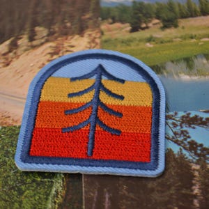 May include: A blue, orange and yellow embroidered patch with a stylised pine tree in the centre. The patch has a rounded top and a straight bottom.