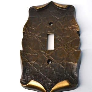 May include: A decorative light switch cover with a textured, bronze-colored finish. The cover has a shield-like shape with a single toggle switch opening and two screw holes. Gold-colored accents are visible along the edges.