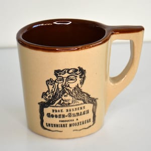May include: Tan ceramic mug with a brown interior and handle. The mug features a black and white illustration of a man with a mustache and the text "Prof. Halbert Goose-Grease Produces a Luxuriant Moustache."