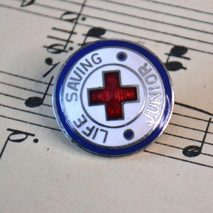 May include: A white and blue enamel pin with a red cross in the center. The pin says "Life Saving Junior" around the outside.