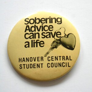 May include: A round, pale yellow pin-back button featuring black text that reads "Sobering Advice can save a life" and "HANOVER CENTRAL STUDENT COUNCIL." A black graphic illustration of a metal key attached to a heart-shaped charm is also present.