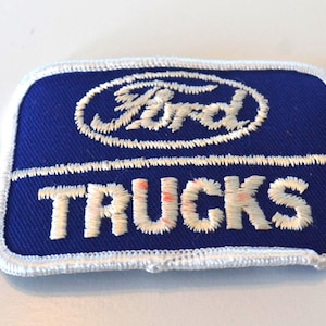 May include: A rectangular blue patch with white embroidered text and border. The word "TRUCKS" is below the Ford logo. The Ford logo and text are white. The patch is likely for a Ford truck.