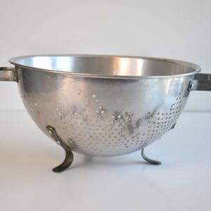 May include: A vintage, silver-toned metal colander with a rounded bowl shape. It features small drainage holes, two side handles, and three curved feet for stability. The kitchenware item is designed for draining liquids from food.