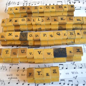 May include: Vintage wooden alphabet stamps arranged on sheet music. The stamps are rectangular with individual letters and numbers printed in black ink. The wood has a warm, aged tone, and the letters are in a classic serif font.