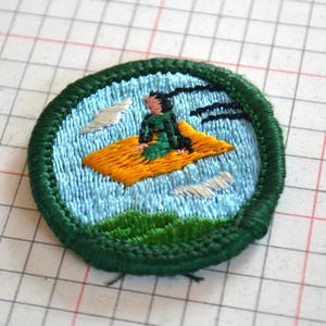 May include: Embroidered patch featuring a person riding a magic carpet. The patch has a green border, a light blue background, and a yellow carpet. The person is wearing green and black clothing. White clouds and green grass are also visible.