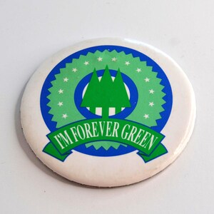 Könnte beinhalten: Ein runder Button mit weißem Hintergrund und grün-blauem Design. Das Design umfasst drei grüne Bäume, einen grünen Ring mit weißen Sternen und einen blauen Ring. Der Text "I'M FOREVER GREEN" ist auf einem grünen Banner zu sehen.