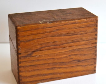 May include: A rectangular wooden box with a natural brown wood grain finish and visible finger joint construction on the corners. The box features a horizontal lid seam across the middle, showcasing a simple, functional design.