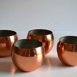 May include: Four copper-coloured, spherical bowls with a dark interior. The bowls have a shiny, reflective surface, and are arranged on a white surface. The bowls are likely for decorative or serving purposes.