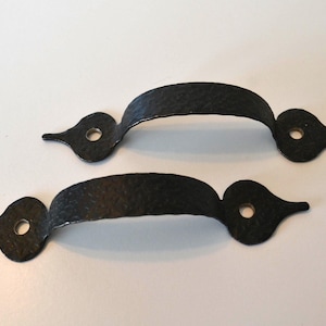 May include: Two black, wrought iron handles with a hammered texture. Each handle has a curved grip and decorative, teardrop-shaped ends with a single hole for mounting. The handles are designed for doors, drawers, or cabinets.