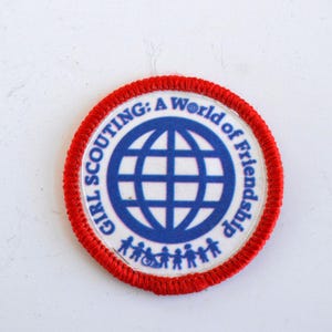 May include: A red and white Girl Scout patch with a blue globe and the text "Girl Scouting: A World of Friendship".