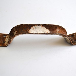 Puede incluir: Un tirador de metal oxidado y arqueado, con placas de montaje en cada extremo. El tirador tiene un diseño curvado con un parche blanco en el centro. El metal muestra signos de edad y desgaste, con óxido marrón y naranja.