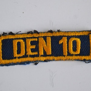May include: A blue and yellow fabric patch with the text "DEN 10" in yellow lettering.