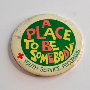 May include: A round, vintage button with the text "A PLACE TO BE SOMEBODY" in red and yellow letters on a green background. The words "YOUTH SERVICE PROGRAMS" are in a ring around the edge. A red cross is on the left side.