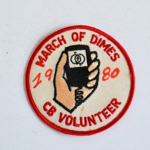 Könnte beinhalten: Ein weißer und roter runder Aufnäher mit dem Text "March of Dimes 1980 CB Volunteer" und einer Hand, die ein schwarz-weißes Gerät mit einem Symbol einer Person in einem Kreis hält.