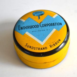 Puede incluir: Una lata amarilla vintage con un borde negro y un diseño azul y blanco en la tapa. La tapa presenta el texto "Underwood Corporation" y "Sundstrand Ribbon". La lata es redonda.