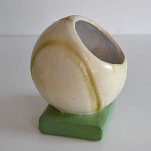 May include: A ceramic baseball-shaped planter or container with a glossy off-white finish and textured yellow stitching details. The object is mounted on a solid, rectangular, matte green base, featuring a wide, angled circular opening at the top.