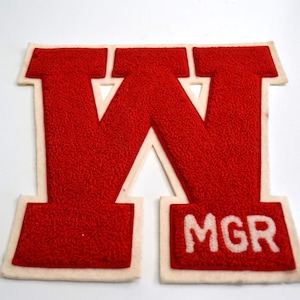 May include: A red felt letter "W" patch with a cream-coloured border. The text "MGR" is embroidered in white at the bottom. The patch is likely for a varsity jacket or similar apparel.