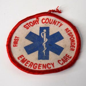 May include: A circular embroidered patch with a white background and a red stitched border. It features a blue Star of Life symbol with a white serpent and staff in the center. The text reads FIRST STORY COUNTY RESPONDER EMERGENCY CARE.