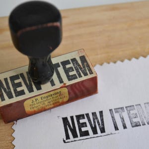 May include: A rectangular rubber stamp with a black wooden handle and a base featuring the text NEW ITEM in bold black letters. A small yellow label on the side displays J.P. Engraving and contact details. The stamp has left a black ink impression.